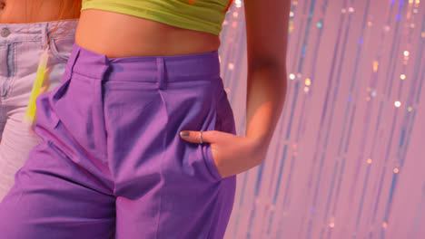 Studio-Close-Up-Of-Two-Women-Showing-Hips-And-Torsos-Standing-In-Front-Of-Tinsel-Curtain-Against-Pink-Background